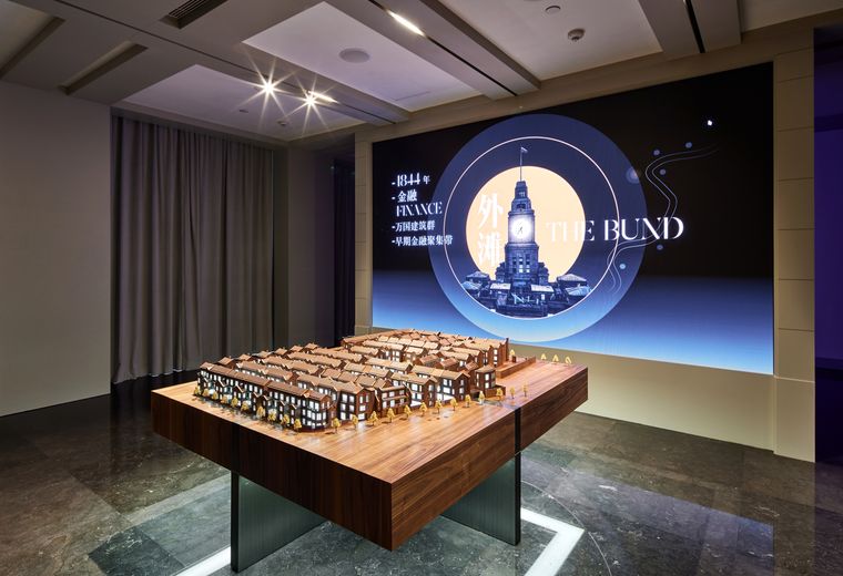Long display table carrying a district model in front of a large screen